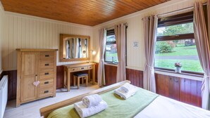 Quarto com teto de madeira, guarda-roupa de madeira, escrivaninha com cadeira, cama com toalhas e janela com cortinas.