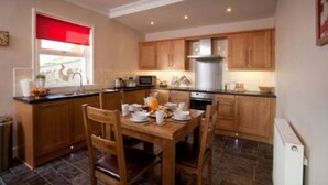 A compact kitchen with wooden cabinets, a dining table with chairs, and a window with a red blind. - Rowan Tree Cottage (Cupar)