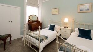 A bedroom with two beds, a dresser, a vanity with a mirror, and a window with a curtain. - Seafield (North Berwick)