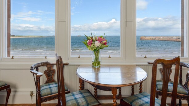 A dining area with a table and chairs, a vase with flowers, and a view of the sea through large windows. - Seafield (North Berwick)