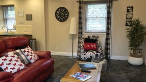 A living room with a red leather sofa, a wooden coffee table with a book and remote, a patterned armchair with a 'Love' pillow, a wall clock, and a potted plant. - Cherry Blossom Cottage (Cupar)