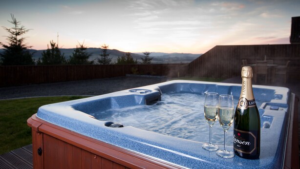 A hot tub with two glasses of champagne and a bottle, set against a scenic backdrop of trees and hills. - Cherry Blossom Cottage (Cupar)