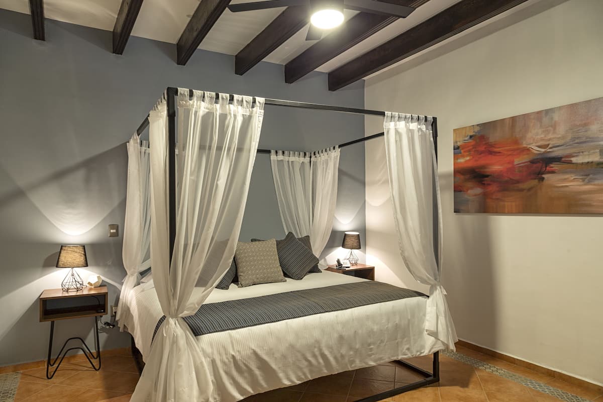 A four-poster bed with white linens and a canopy, a bedside table with a lamp, and a painting on the wall.