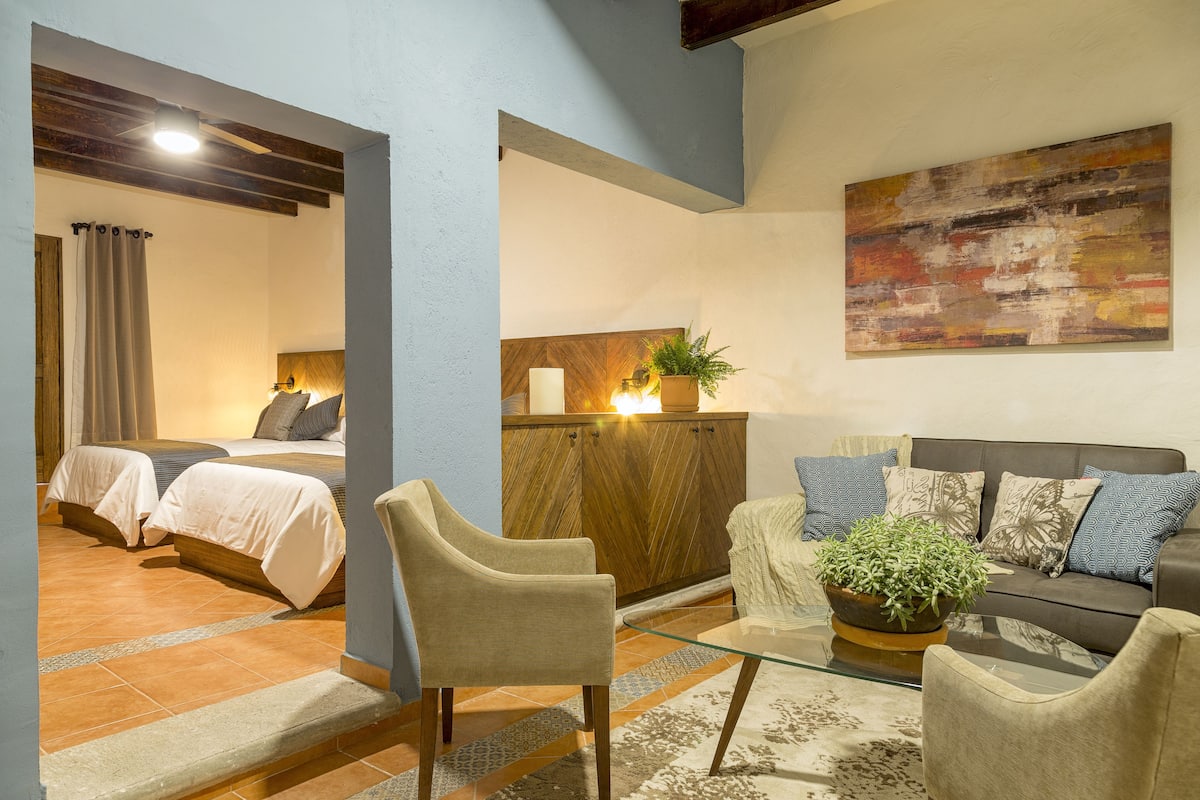 A hotel room with two beds, a sofa, a glass coffee table, and a potted plant.