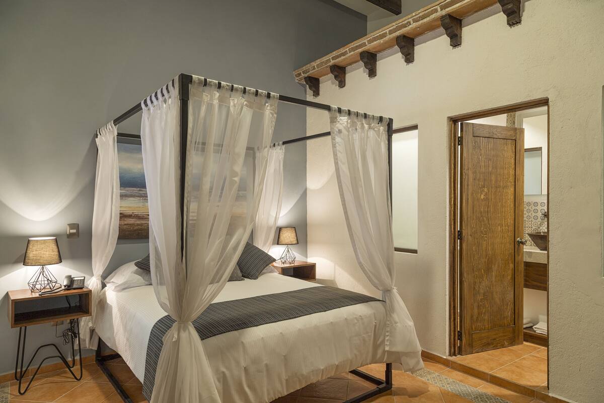 A four-poster bed with sheer curtains, bedside lamps, and a view of the outdoors.