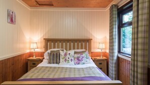 A bedroom with a wooden headboard, two bedside tables with lamps, a bed with floral bedding, and a window with plaid curtains. - Bluebell Lodge 2 with Hot Tub (Newton Stewart)