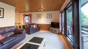 A cozy living room with a brown leather sofa, a glass-top coffee table, and a television. There are framed pictures on the wall and a dining area with a table and chairs. - Bluebell Lodge 2 with Hot Tub (Newton Stewart)