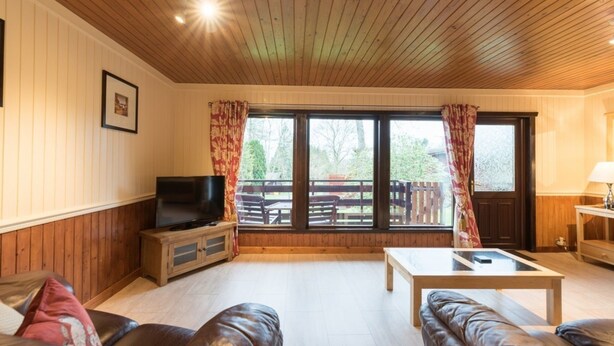 A living room with a wooden ceiling, a television, a sofa, a coffee table, and a view of a deck and trees. - Bluebell Lodge 2 with Hot Tub (Newton Stewart)