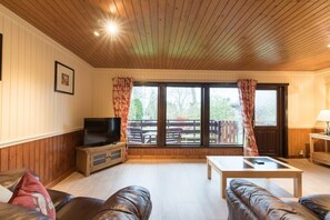 Una sala de estar con techo de madera, televisor, sofá, mesa de centro y vista a una terraza y árboles.