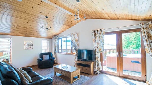 Ein Wohnzimmer mit Holzbalkendecke, einem schwarzen Ledersofa, einem Couchtisch aus Holz, einem Flachbildfernseher auf einem Holzständer und einer Glastür zum Außenbereich.