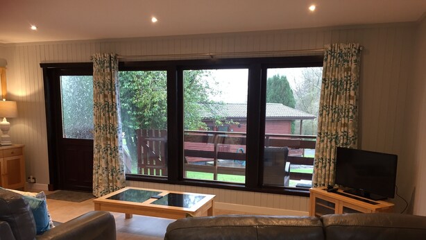 A living room with a sofa, a coffee table, a TV on a stand, and large windows with curtains.