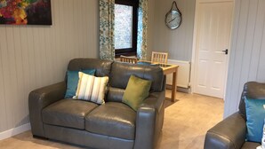 A living room with a brown leather sofa, a dining table with chairs, a clock on the wall, and a door.