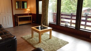 A living room with a wooden floor, a black leather sofa, a wooden coffee table with a decorative item, a lamp, and a view of a wooden deck and trees through large windows.