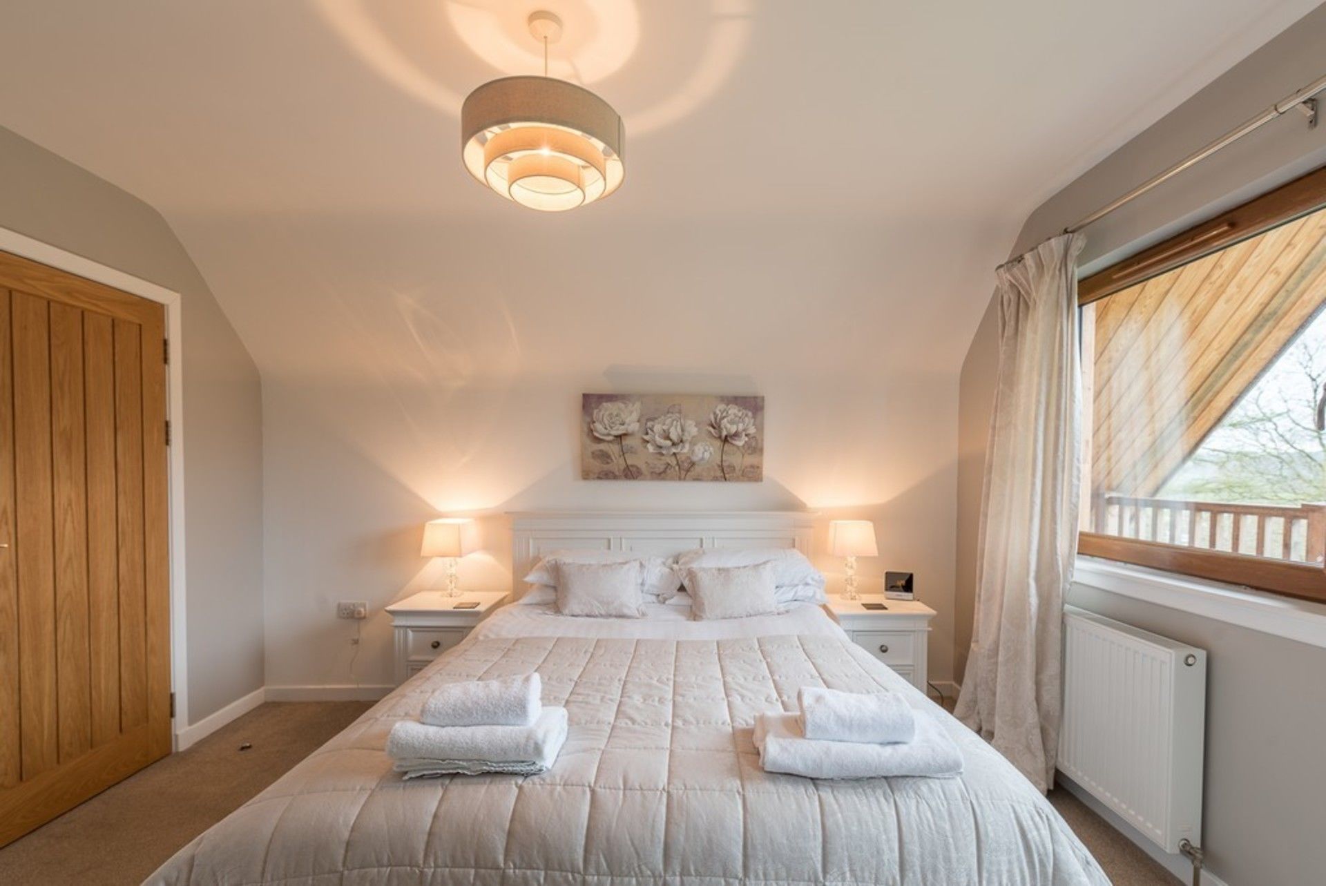 Un dormitorio con una cama grande, dos mesitas de noche, una lámpara en el techo, una ventana con cortinas y una puerta de madera.