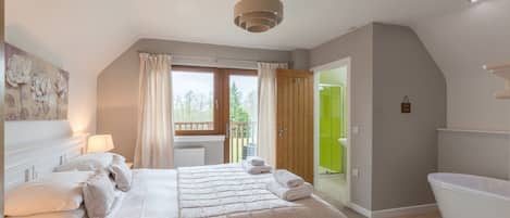 A bedroom with a large bed, a bathroom with a bathtub, and a balcony with a view of trees.
