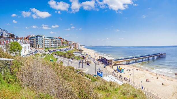 A coastal town with a pier extending into the sea, a beach with people, and a mix of residential and commercial buildings. - Flat 8 Palm Court (Bournemouth)