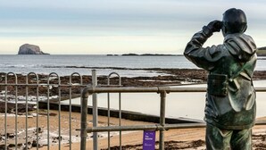 Una estatua de una persona con binoculares contemplando una playa con costa rocosa y una isla distante.