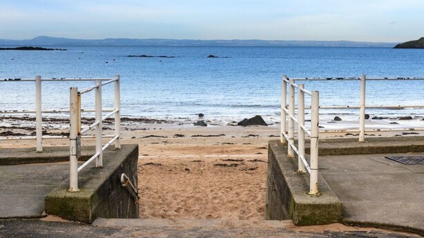 Una escalera de concreto que desciende hacia una playa de arena, con barandillas a ambos lados, con vista a un mar tranquilo y tierra distante.