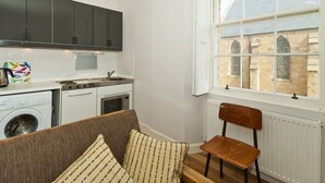 Una cocina compacta con electrodomésticos modernos, un ventanal con vistas a edificios históricos y una zona de estar acogedora con un sofá estampado y una silla de madera.