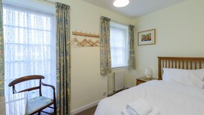 A hotel room with a bed, a chair, a coat rack, and a window with curtains. - Swifts Cottage (Tranent)
