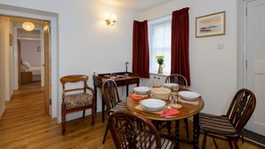 A dining area with a round table set for four, surrounded by wooden chairs. There is a window with red curtains, a wall-mounted light fixture, and a door leading to another room. - Swifts Cottage (Tranent)