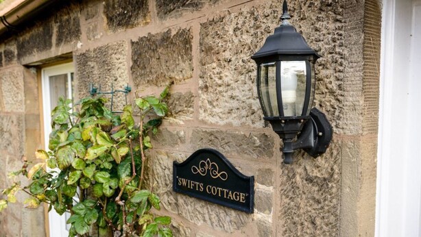 A stone wall with a 'SWIFTS COTTAGE' sign and a black wall-mounted lantern. - Swifts Cottage (Tranent)