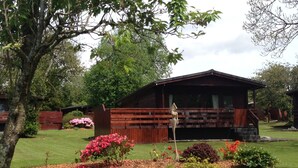 A wooden cabin with a fenced yard, surrounded by trees and flowering plants. - Bluebell Lodge 3 with Hot Tub (Newton Stewart)