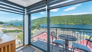 A balcony with a view of a lake and mountains, a table, and two chairs.
