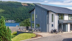 A modern two-story building with a balcony overlooking a lake, surrounded by forested hills.