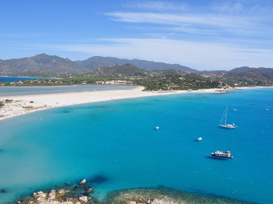 A coastal landscape with a beach, clear turquoise waters, and boats. Mountains are visible in the background.