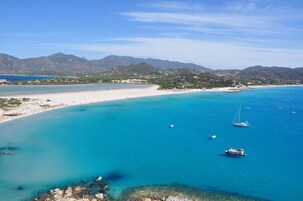A coastal landscape with a beach, clear turquoise waters, and boats. Mountains are visible in the background.