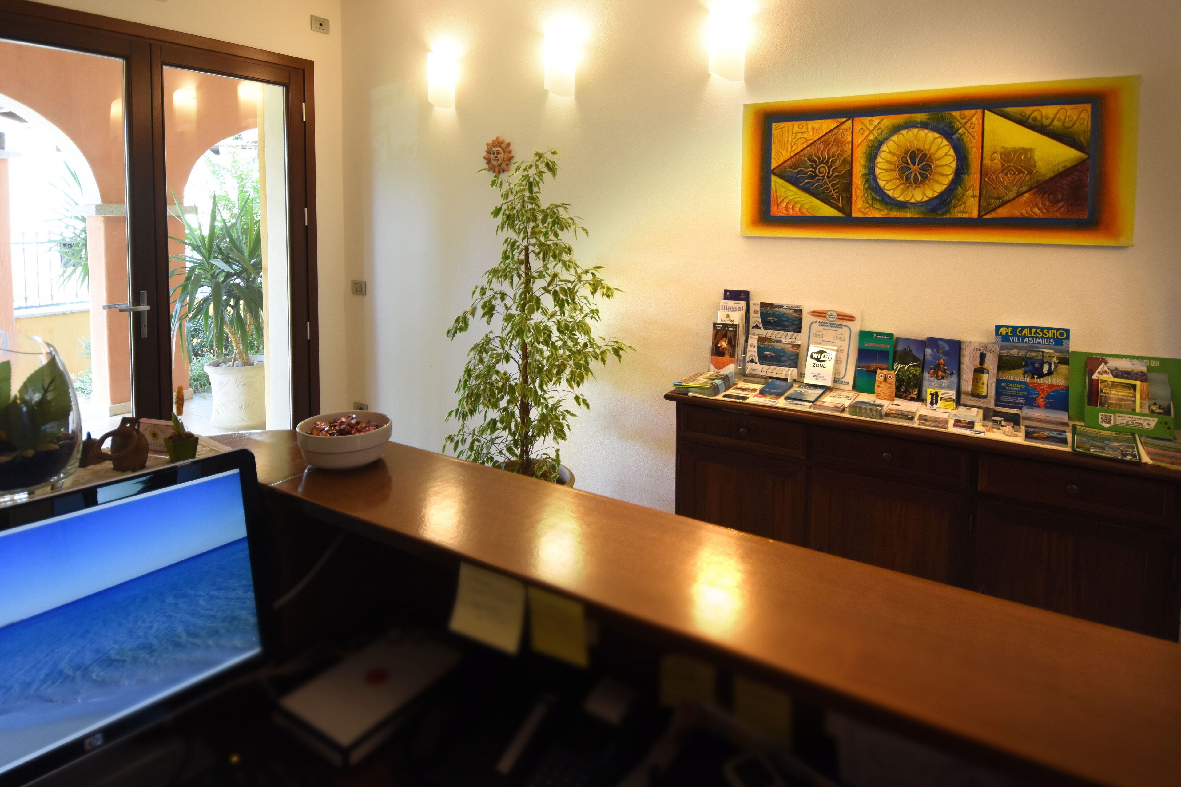 A reception desk with a laptop, a potted plant, and a wall with a framed artwork.