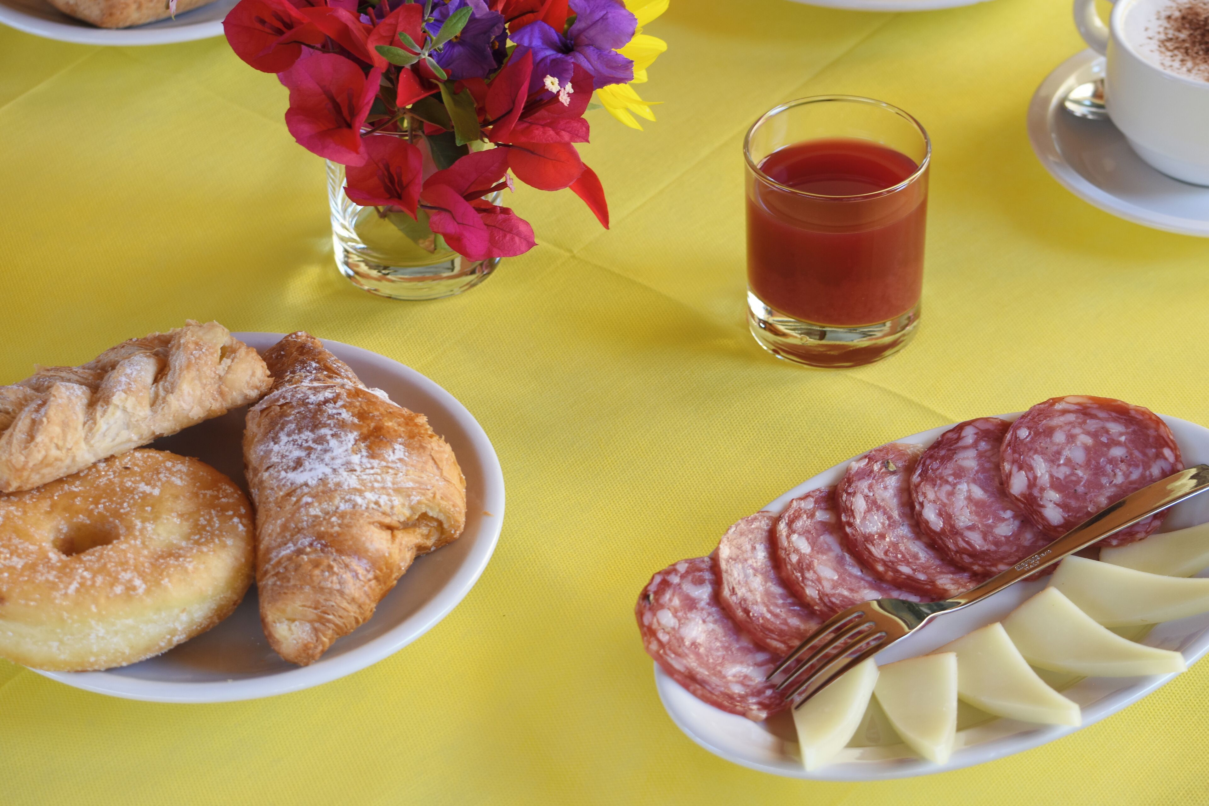 Una mesa con mantel amarillo, un plato de pasteles, un vaso de jugo rojo y un plato de salami en rodajas.
