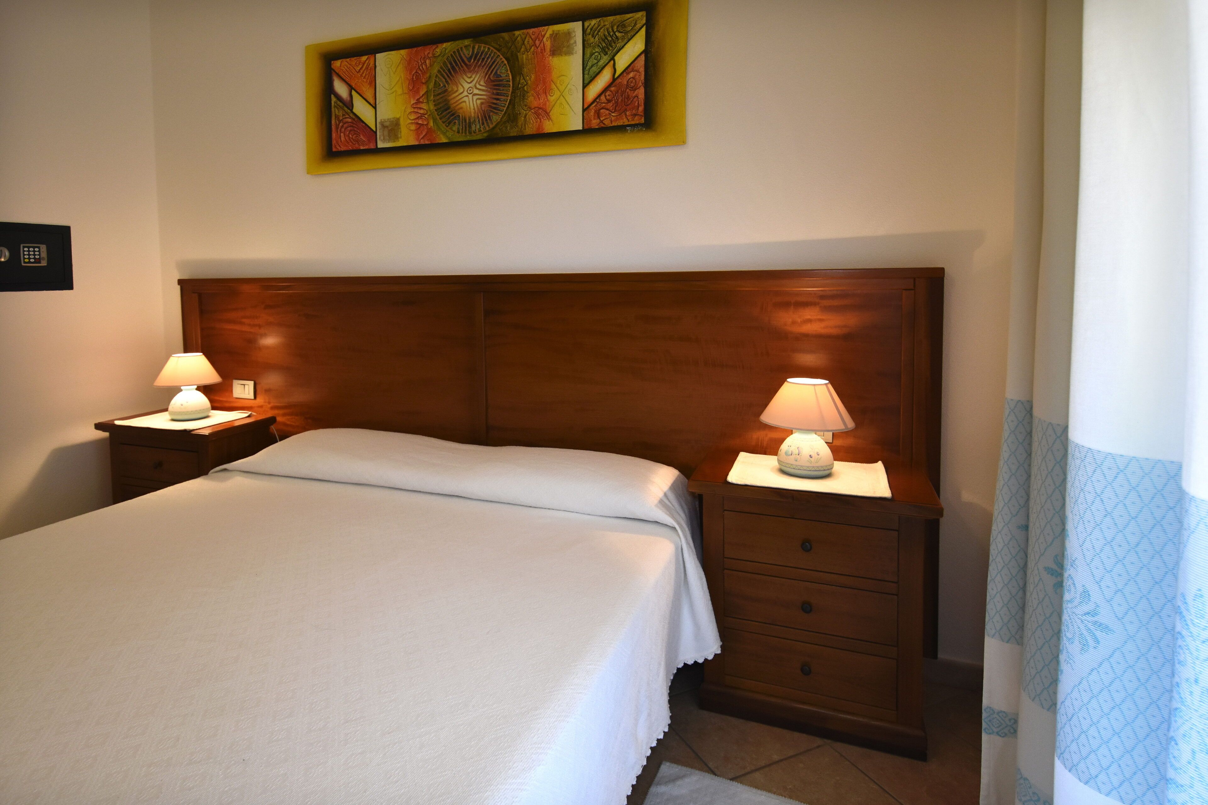 A neatly made bed with white linens, two bedside tables with lamps, a wooden headboard, and a colorful abstract painting above the bed.