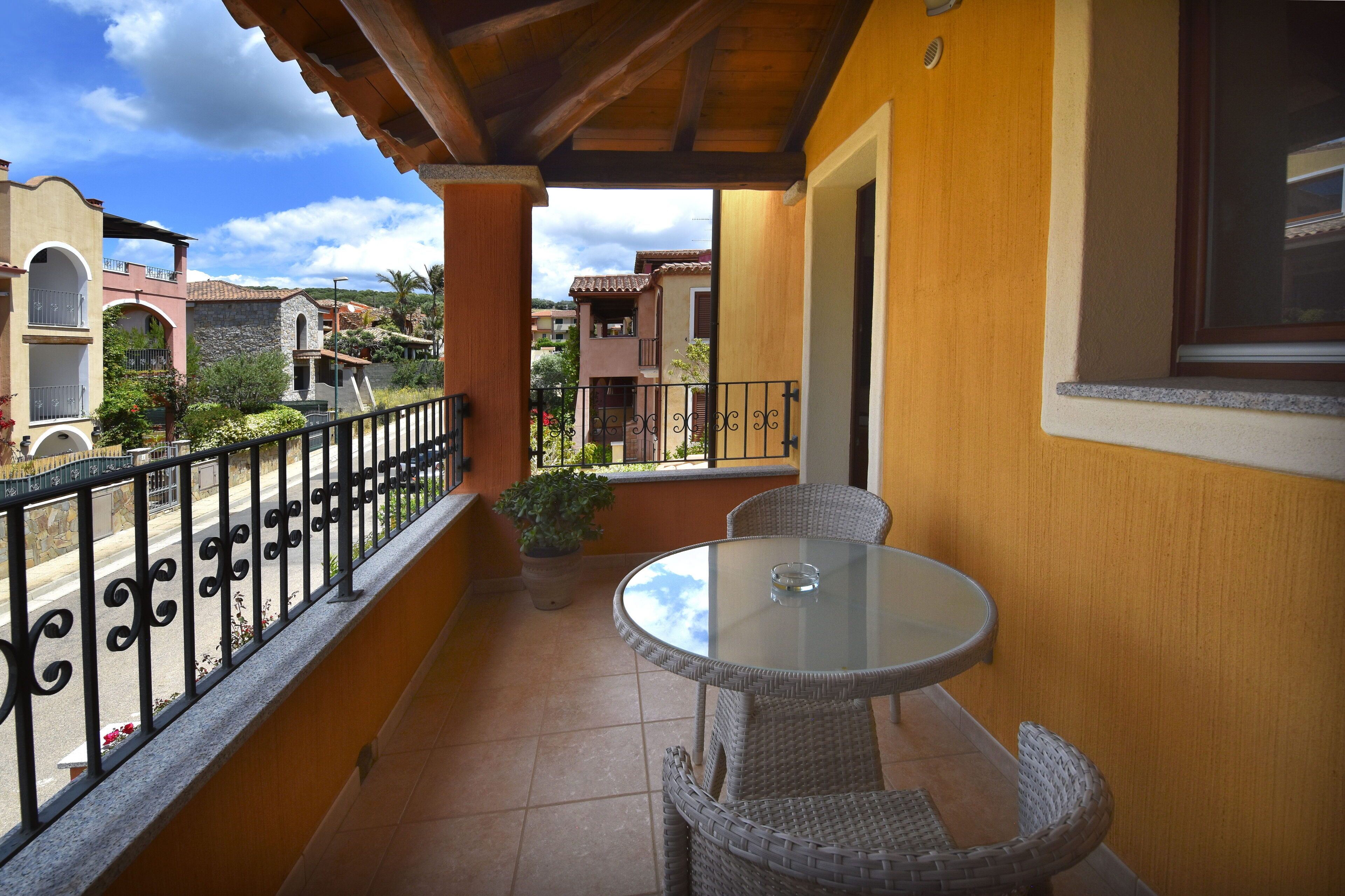 A balcony with a round table and wicker chairs, overlooking a street with buildings and a clear sky.
