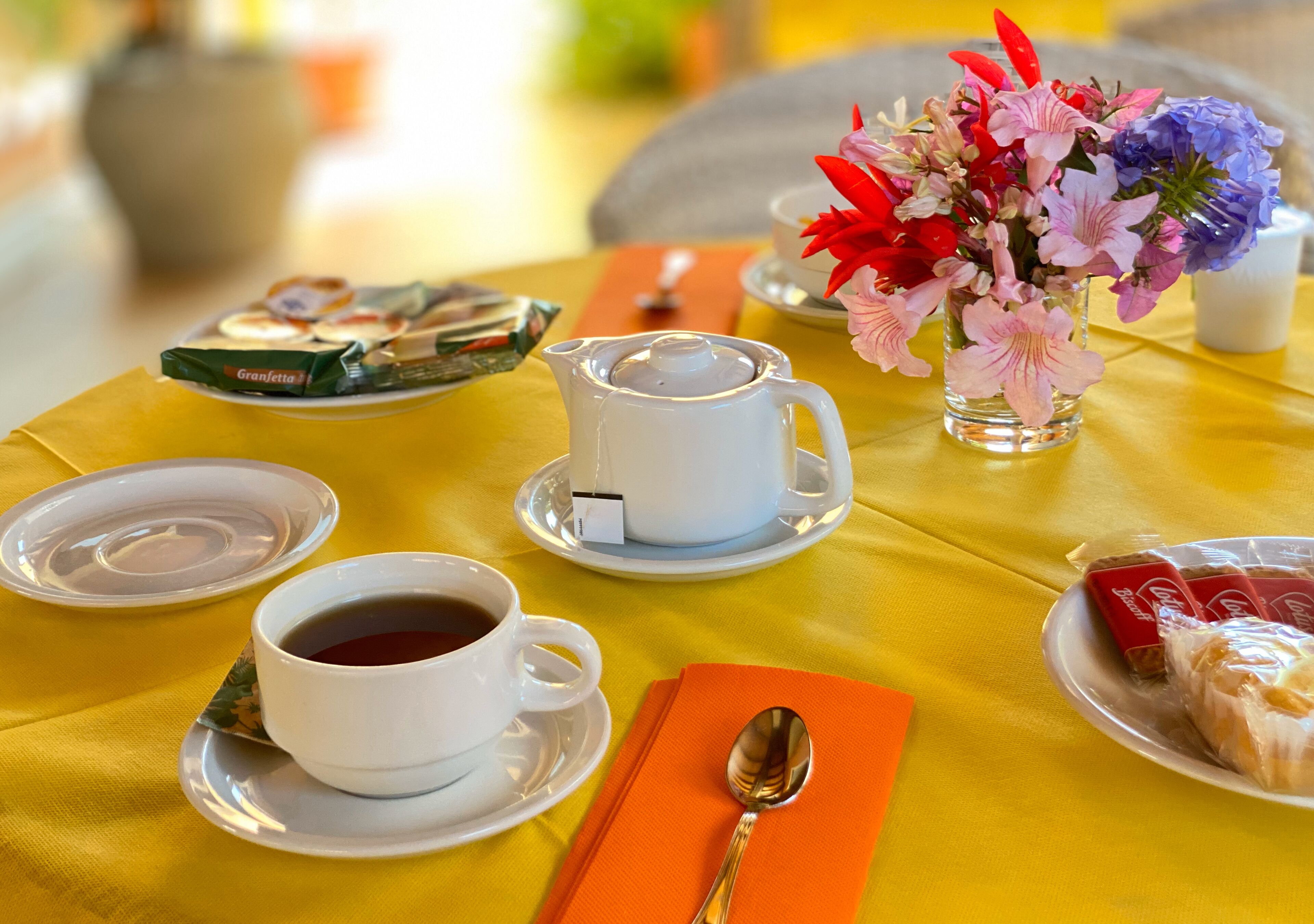 Una mesa servida con tetera blanca, dos tazas de té, un jarrón con flores, un plato de pasteles y una cuchara sobre un mantel naranja.