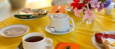 A table set with a white teapot, two cups of tea, a floral vase, a plate with pastries, and a spoon on an orange napkin.