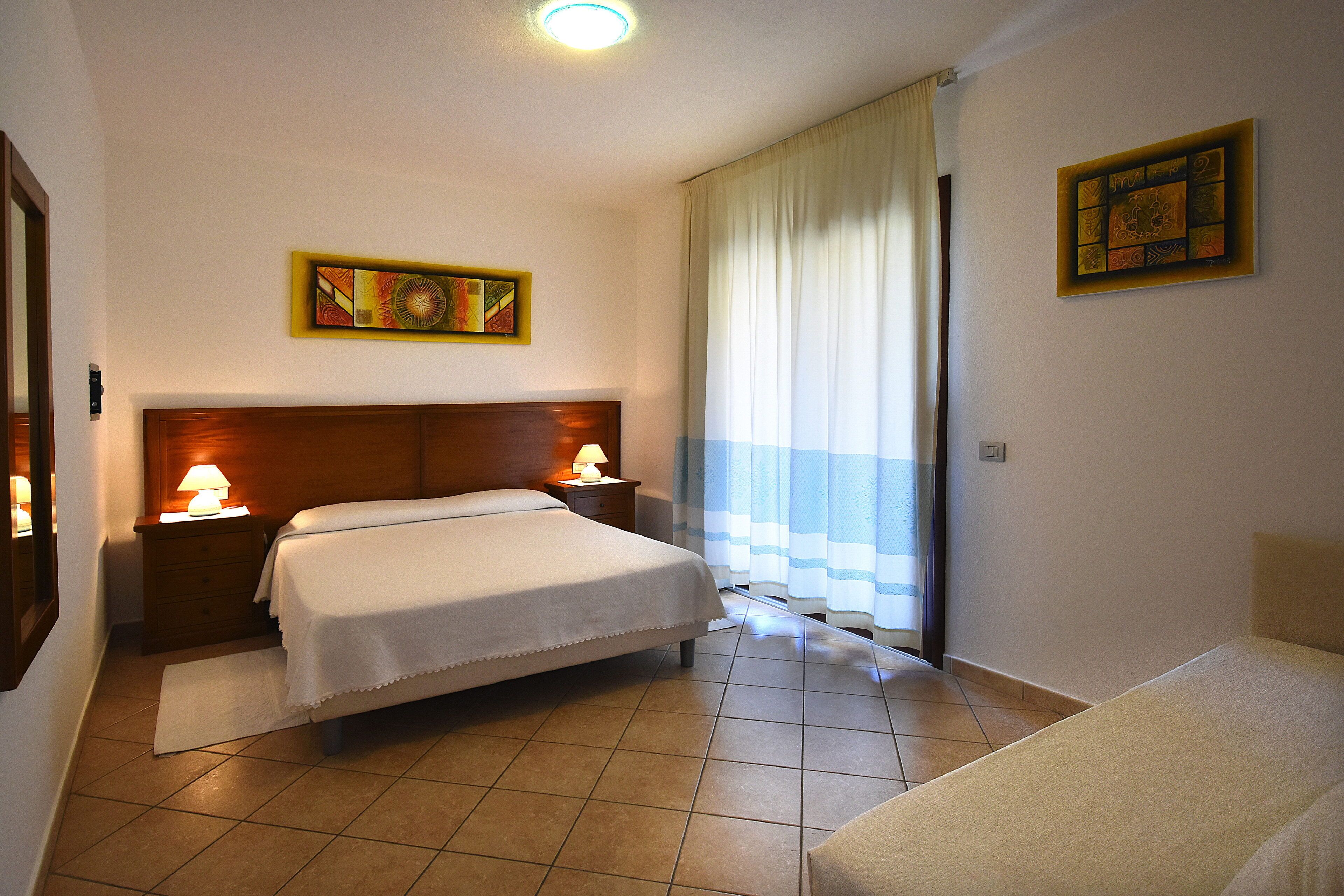 A hotel room with a bed, bedside tables, a lamp, a window with curtains, and a painting on the wall.