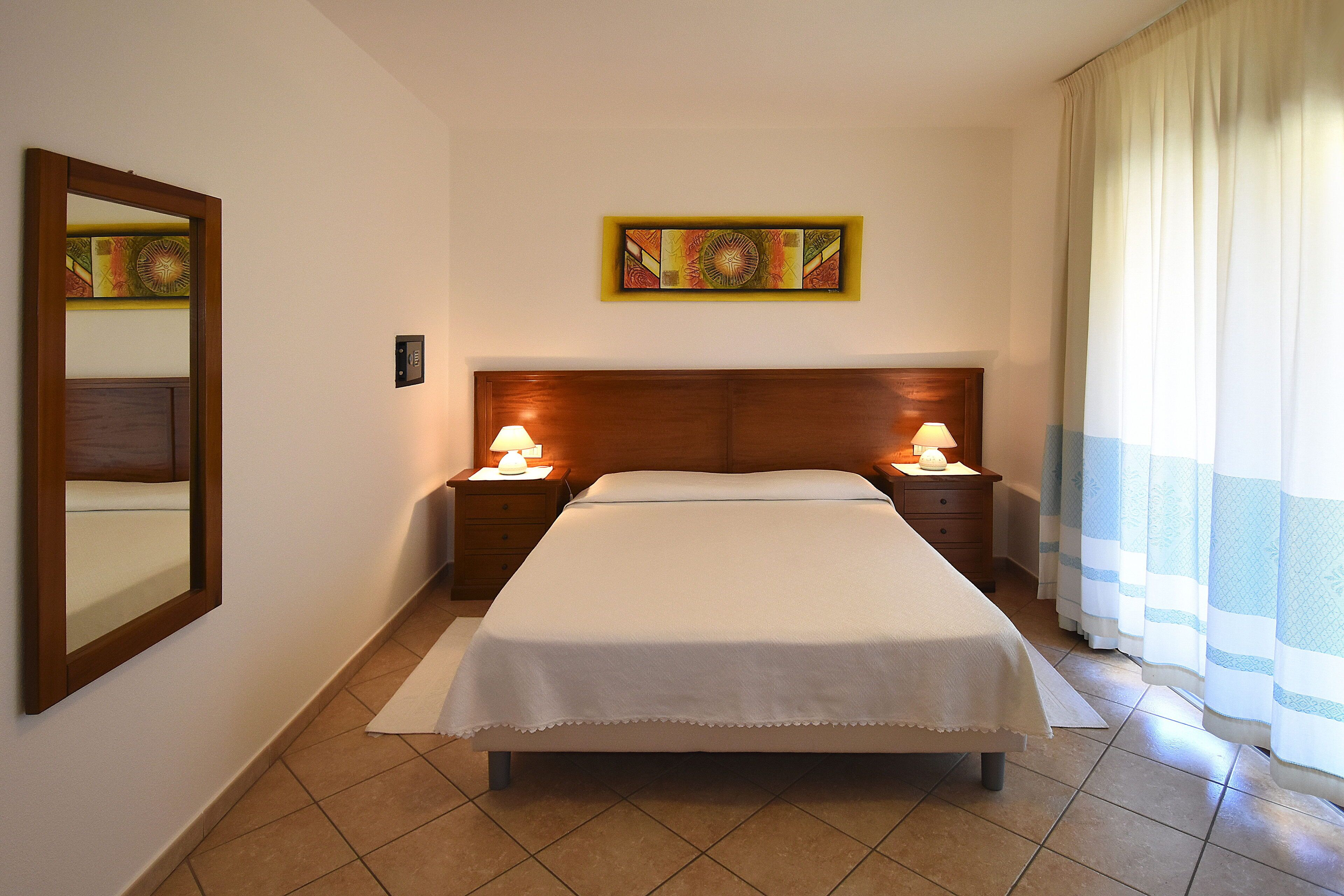 A hotel room with a bed, two bedside tables with lamps, a wall-mounted artwork, a mirror, and a window with curtains.