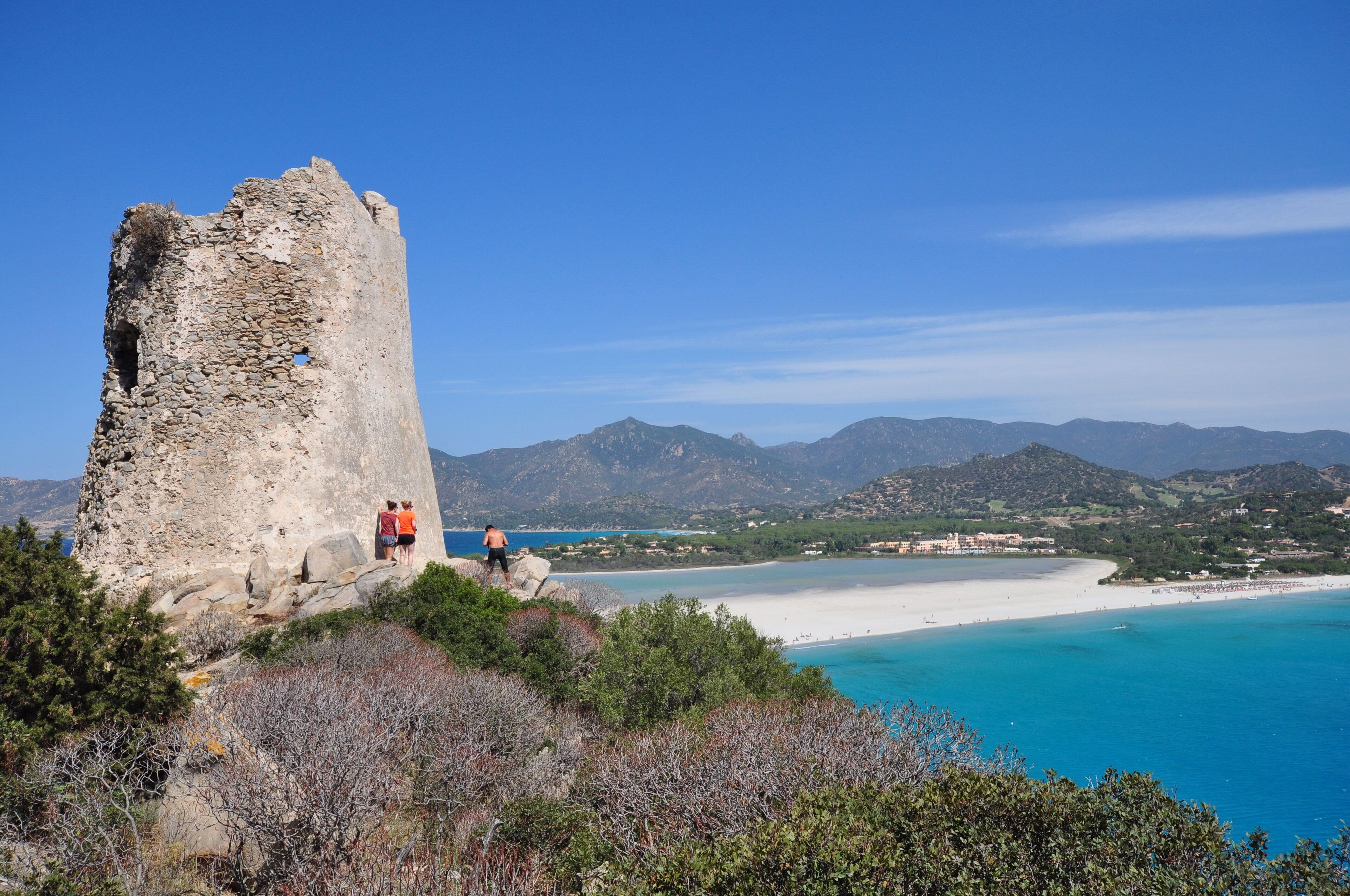 Paisaje costero con una torre de piedra histórica, una playa y terreno montañoso al fondo.
