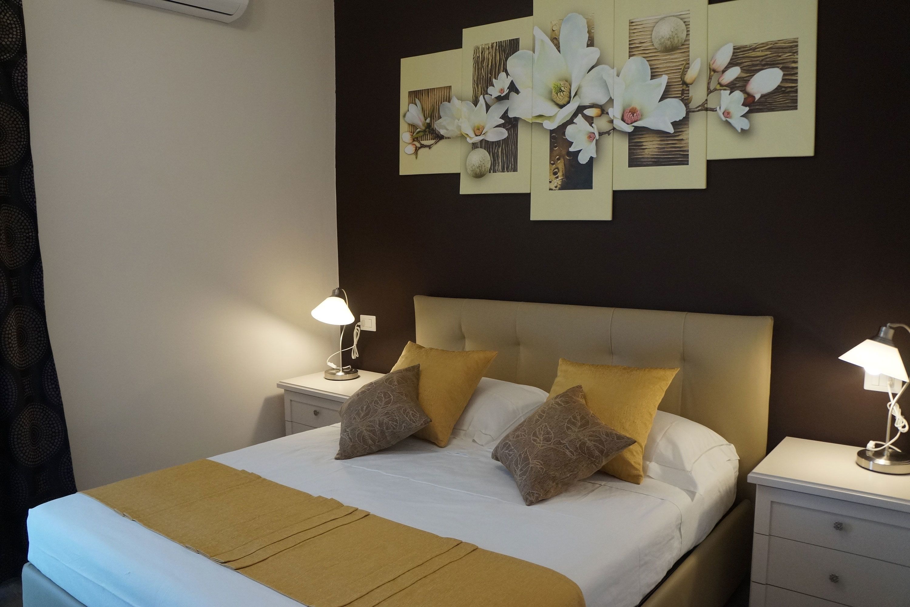 Een net opgemaakt bed met een beige hoofdbord, twee nachtkastjes met lampen, en een kunstwerk erboven.