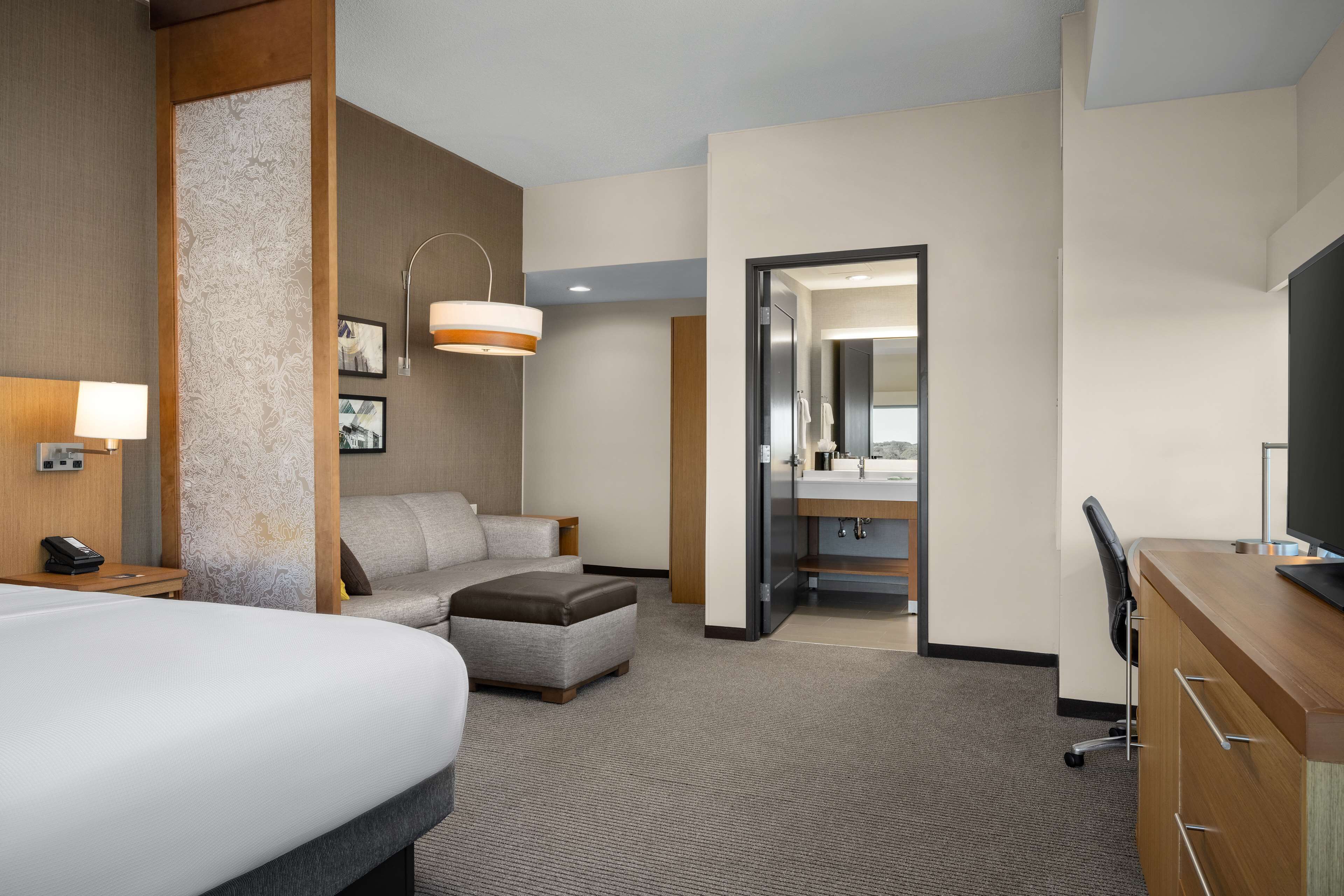 A hotel room with a large bed, a sofa, a desk, a chair, a lamp, a mirror, and a bathroom visible through an open door.
