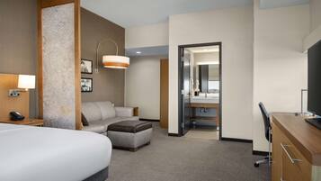 A hotel room with a large bed, a sofa, a desk, a chair, a lamp, a mirror, and a bathroom visible through an open door.