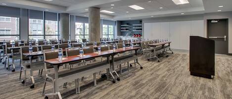 A conference room with multiple tables and chairs, equipped with water bottles, in a modern building with large windows and a carpeted floor.
