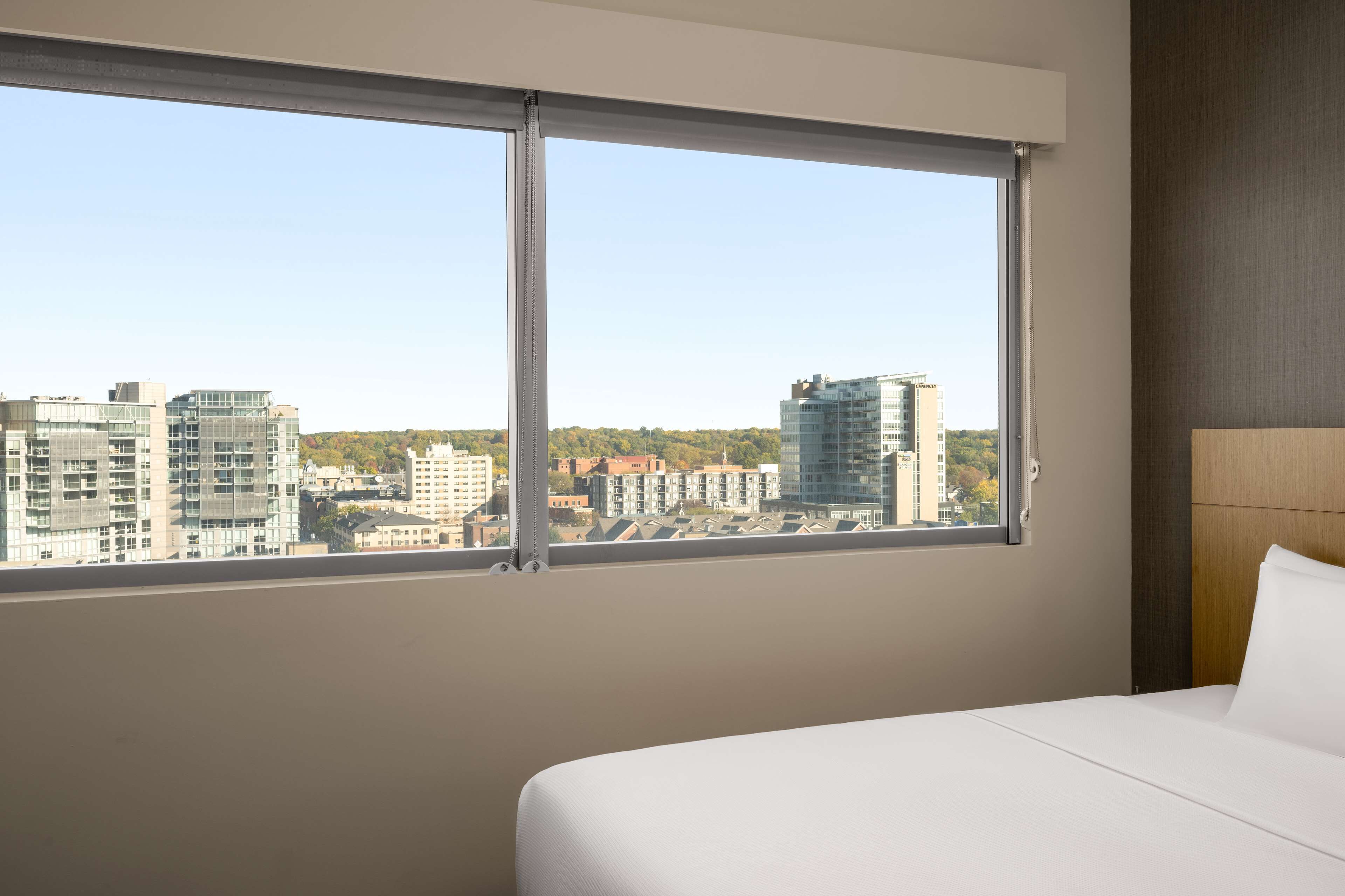 Habitación de hotel con un ventanal grande que ofrece vistas a la ciudad, una cama bien hecha y un cabecero moderno.