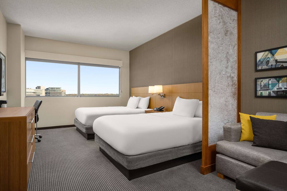 Une chambre d’hôtel avec deux lits, un bureau, une chaise, un canapé et une grande fenêtre donnant sur la ville.
