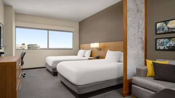 Quarto de hotel com duas camas, uma escrivaninha, uma cadeira, um sofá e uma janela grande com vista para a cidade.