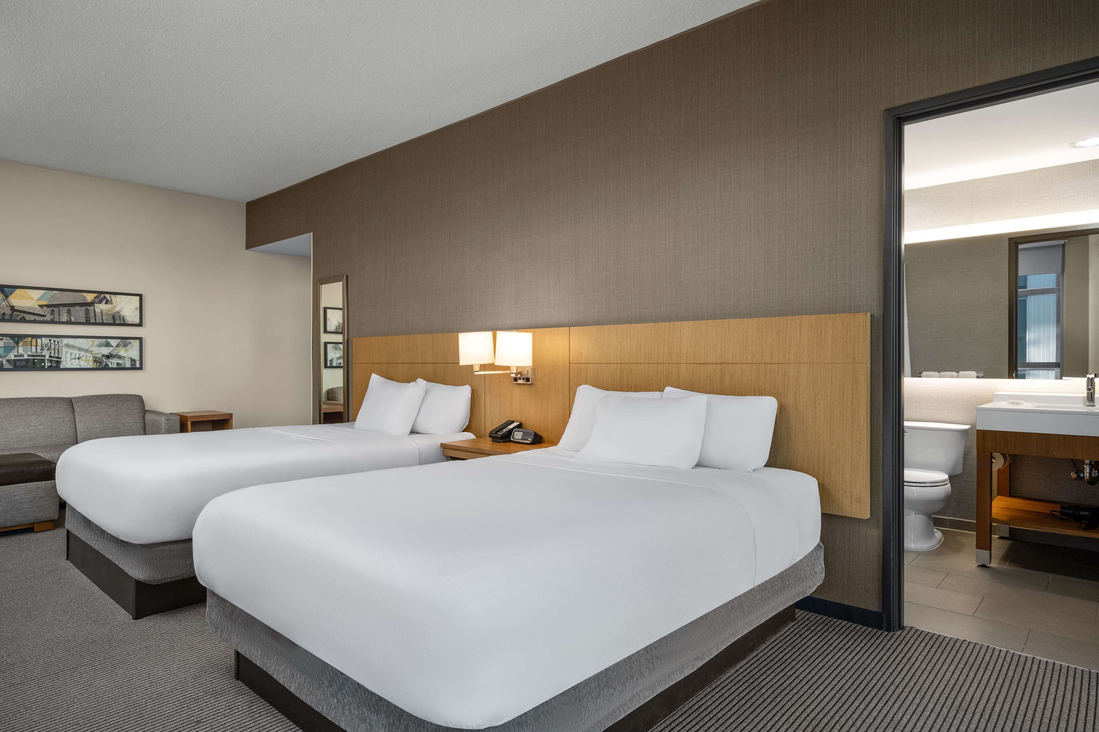 A hotel room with two beds, a sofa, a bedside table with a lamp, and a bathroom visible through an open door.