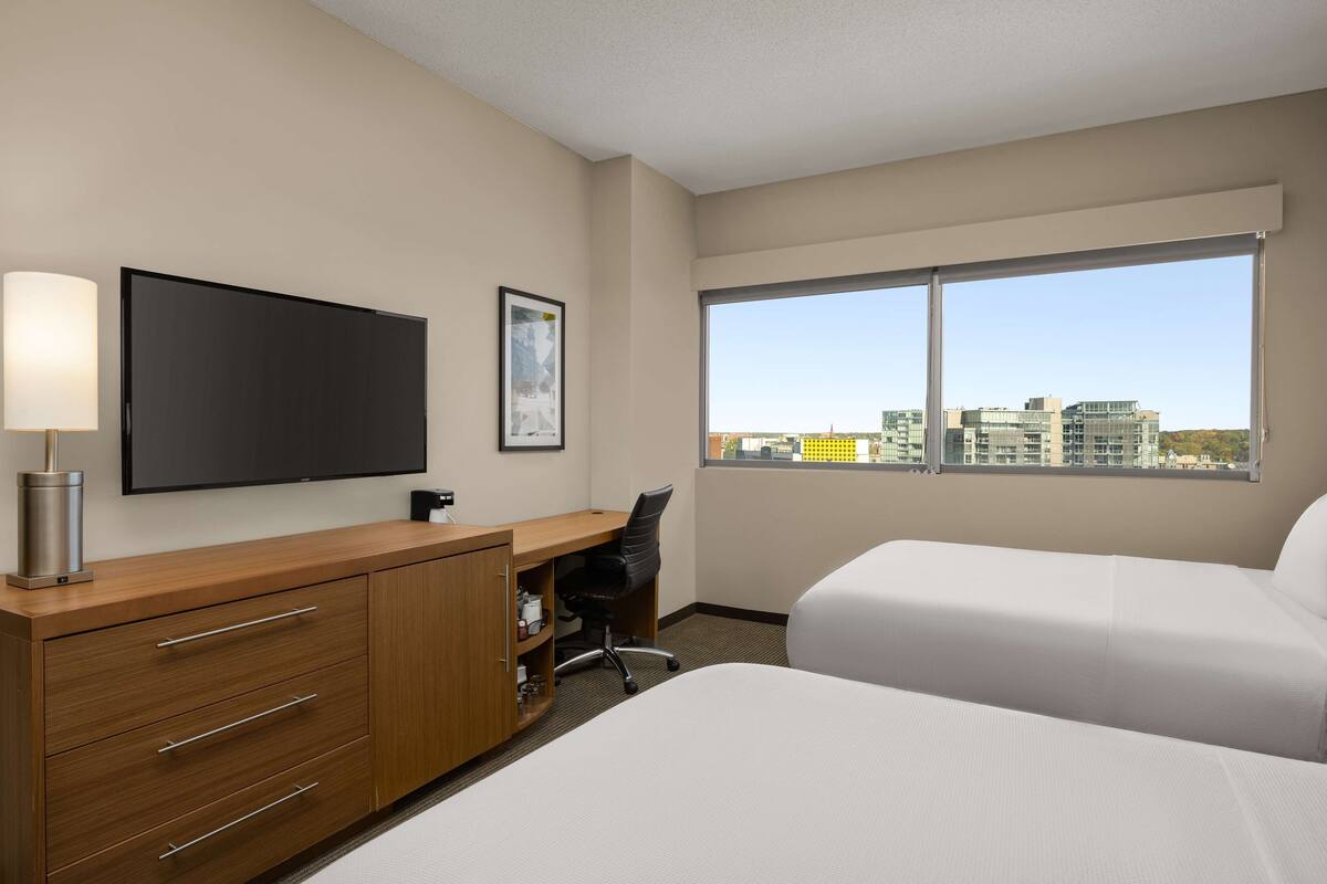 Une chambre d’hôtel comprenant un lit, un bureau avec une chaise, une télévision et une grande fenêtre donnant sur la ville.