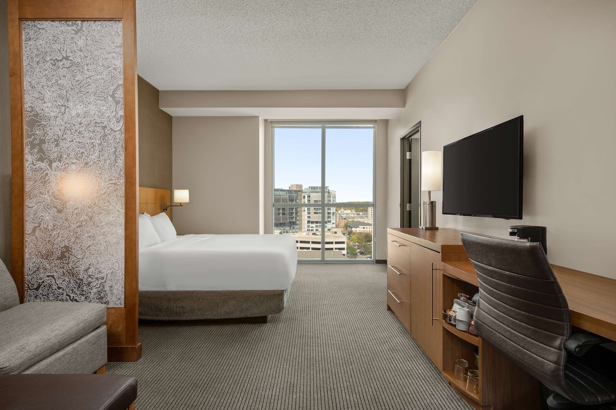 Une chambre d’hôtel dotée d’un grand lit, d’un bureau avec un téléviseur à écran plat, d’une chaise, d’une commode et d’une grande fenêtre donnant sur la ville.